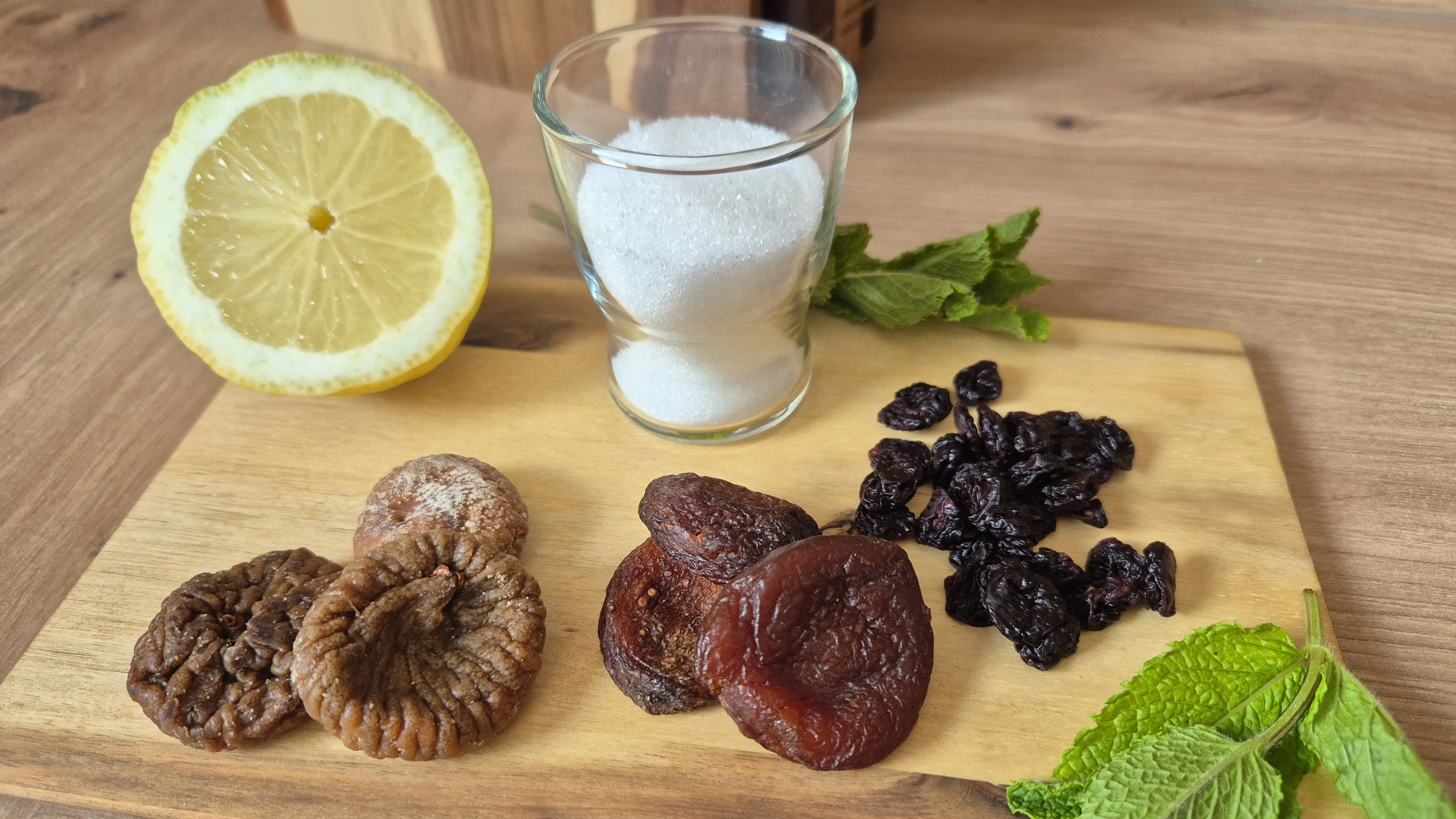 Zutaten für einen Wasserkefir-Ansatz auf einem Holzbrett: halbe Zitrone, ein Glas Zucker, getrocknete Feigen, Aprikosen, Rosinen und Minze